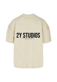 Bežs liela izmēra t-krekls ar tekstu "2Y STUDIOS" trešajā daļā biezā melnā fontā, ar papildu tekstu zem tā rakstītā rokrakstā.