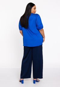 Blouse bleue avec des manches bouffantes, associée à un pantalon large bleu marine et des sandales à talons hauts bleues. Tissu lisse et design décontracté.