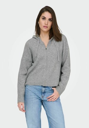 Sweat à capuche zippé gris avec ourlet et poignets côtelés, présentant une coupe décontractée et des cordons de serrage, associé à un jean bleu.