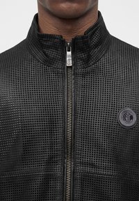 Veste noire texturée avec une fermeture éclair centrale argentée et un patch rond avec logo sur la poitrine gauche portée par une personne.