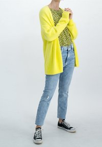 Gelber flauschiger Cardigan über einem schwarzen Blumenhemd, kombiniert mit hellblauen, ausgefransten Jeans und schwarzen Turnschuhen. Lässiges, geschichtetes Outfit mit hellen Farben.