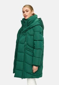Piumino verde con colletto alto e ampio cappuccio. Presenta sezioni trapuntate, tasche frontali con zip e un design dal taglio rilassato.