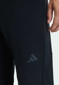 Čierne športové nohavice z hladkej tkaniny, s zužujúcimi sa nohavicami a vyrazeným logom na stehne. Švové detaily zdôrazňujú štruktúru.