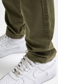 Olijfgroene katoenen broek met een gestructureerde afwerking, gedragen met witte leren sneakers met perforaties en grijze accenten.