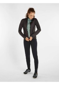 Giacca sportiva nera sopra una maglietta azzurra chiara, abbinata a leggings scuri e calzature grigie. Tessuto liscio, design aderente, branding minimale.