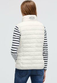 Weißer gesteppter Weste mit hohem Kragen und Logo auf dem Rücken, getragen über einem schwarz-weißen gestreiften Langarmshirt, kombiniert mit dunklen Jeans.