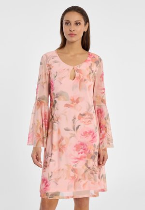 Frau trägt ein rosa, knielanges Kleid mit Blumenmuster, Glockenärmeln und kleinem Schlüssellochausschnitt vorne, steht vor einem schlichten weißen Hintergrund.