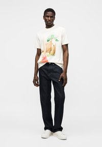 Mies, jolla valkoinen graafinen t-paita, tummansiniset farkut ja valkoiset lenkkarit, seisoo yksinkertaisen vaalean taustan edessä.