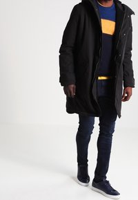 Mann in schwarzem Langmantel, gestreiftem blauen und gelben Pullover, dunklen Jeans und marineblauen Sneakers, der vor einem weißen Hintergrund läuft.