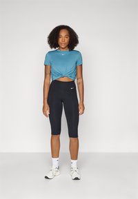 T-shirt turquoise noué à manches courtes avec un petit logo, associé à des leggings capri noirs. Des baskets blanches et des chaussettes complètent le look.