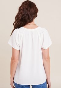 Weiße Kurzarmbluse mit Rundhalsausschnitt und elastischen Details an den Schultern, kombiniert mit gemusterten blauen Hosen.