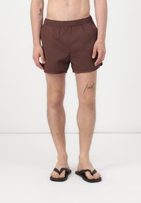 Brune badebukser med elastisk talje, med en glat tekstur. Båret med sorte flip-flops, vist mod en ensfarvet baggrund.