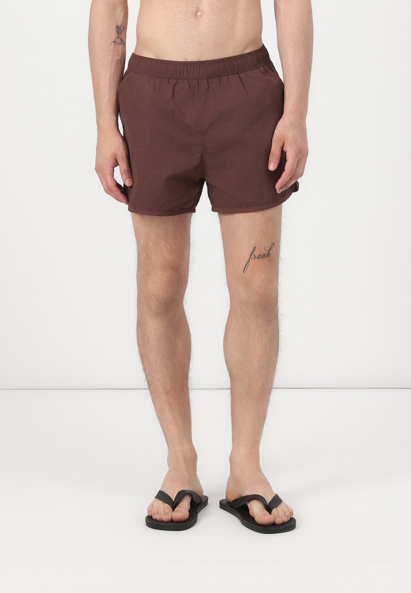 Brune badebukser med elastisk talje, med en glat tekstur. Båret med sorte flip-flops, vist mod en ensfarvet baggrund.