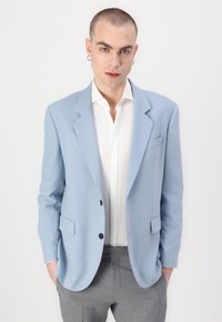 Blazer azul claro de tela suave con solapas de muesca, dos bolsillos frontales y dos botones. Lleva puesto una camisa blanca y pantalones grises.