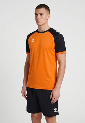 Mann steht, trägt ein orange-schwarzes Sportshirt und schwarze Shorts vor einem schlichten hellen Hintergrund, leicht nach links gewandt.