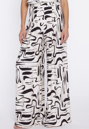 Kobieta nosząca spodnie z wysokim stanem i szerokimi nogawkami z czarnym abstrakcyjnym wzorem na białej tkaninie, stojąca na jednolitym tle.