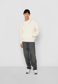 Sweatshirt à capuche crème zippé, coupe décontractée, associé à un jean bleu. Bonnet noir avec un petit patch. Des baskets neutres complètent la tenue.
