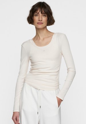 Top lungo beige chiaro a maniche lunghe con una texture a costine e scollatura a girocollo. Presenta una vestibilità aderente e un branding sottile sul tessuto.