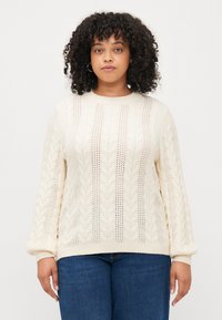 Cream cable-knit sweater with open detailing, round neckline, and long, ribbed sleeves. Worn over blue jeans.