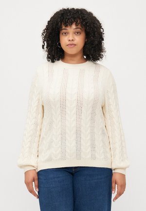 Cream cable-knit sweater with open detailing, round neckline, and long, ribbed sleeves. Worn over blue jeans.