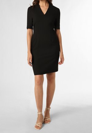 Kobieta w czarnej sukience do kolan z krótkimi rękawami i dekoltem w serek, w połączeniu z beżowymi sandałkami na wysokim obcasie na jednolitym tle.