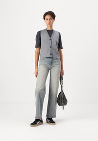 Gilet grigio senza maniche sopra una maglietta nera a maniche corte, abbinato a jeans a gamba larga blu chiaro e sneakers nere con strisce bianche.