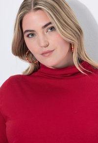 Donna bionda con occhi verdi che indossa un dolcevita rosso e grandi orecchini a cerchio colorati, che guarda leggermente di lato su uno sfondo bianco.