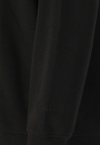 Musta collegepaita, jossa on tekstuuri ja pehmeä kangas. Varressa on ribbireunat ja helma. Merkin nimi "ESSENTIAL" on huomaamaton brodeeraus hihassa.