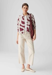 Giacca a camicia a motivo bordeaux e bianco, pantaloni beige e décolleté crema. L'outfit presenta una vestibilità rilassata e un design minimalista.