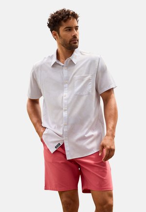 Hombre con cabello rizado, viste una camisa clara de manga corta abotonada y pantalones cortos color coral, de pie con una mano en el bolsillo, mirando hacia un lado.