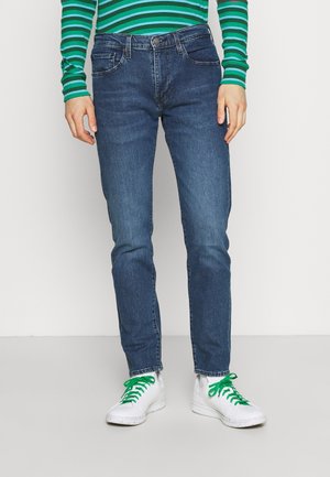 Dunkelblaue Jeans im Slim-Fit mit fünf Taschen und dezenten Stitching-Details. Kombiniert mit weißen Sneakers mit grünen Schnürsenkeln.