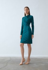 Teal kjole med lange ermer, figursydd silhuett og drapert detaljering ved halsen. Brukt med sandaler med klare hæler på et nøytralt gulv.