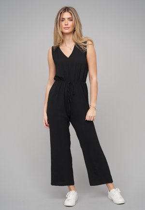 Frau mit langen blonden Haaren trägt einen schwarzen ärmellosen Jumpsuit mit Gürtel, weiße Turnschuhe und minimalistischen Schmuck, steht vor grauem Hintergrund.