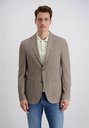 Homme portant un blazer taupe sur un polo crème et un jean bleu, debout devant un fond clair uni.