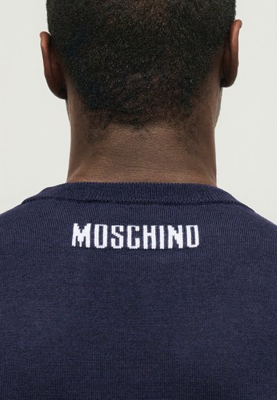 Πίσω όψη ενός ατόμου που φοράει μπλε σκούρο πουλόβερ με τη λέξη "MOSCHINO" σε λευκά πλεκτά γράμματα στην πάνω πλάτη.