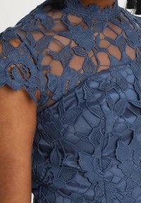 Marinblå spetsklänning med blommönster, genomskinlig halsringning och korta ärmar över ett slätt tyg, buren på övre delen av kroppen.