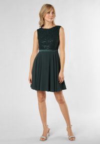 Green A-line dress with a lace bodice, sequin detailing, a pleated skirt, and a satin waistband. Complemented by silver high-heeled sandals.
