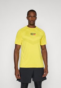 Gul atletisk T-skjorte med korte ermer, rund hals og strukturell design. Fronten viser "BERLIN RUN" i svart og rødt.