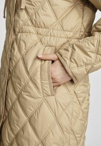 Beżowa pikowana kurtka puchowa z diamentowym wzorem, z boczną kieszenią i teksturowanym wykończeniem. Miękki materiał z matowym wykończeniem.