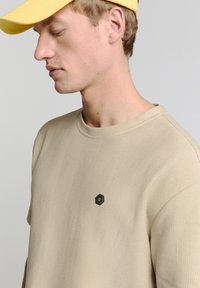 Beige textuurtrui met een hexagonaal zwart logo op de borst, gecombineerd met een gele pet, met een ronde hals en een losse pasvorm.