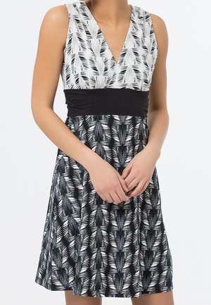 Šaty bez rukávů s černobílými listovými motivy, hlubokým výstřihem do V a širokým černým pásem v pase, oblečené osobou stojící se sepjatýma rukama.