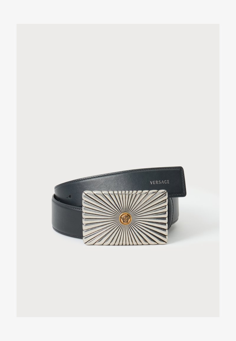 Ceinture en cuir noir avec une boucle rectangulaire argentée ornée de lignes rayonnantes et un petit emblème doré représentant une tête de Méduse au centre.