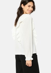 White blouse with ruffled detailing along shoulders and sleeves, made of textured fabric. Features long sleeves and elastic cuffs.