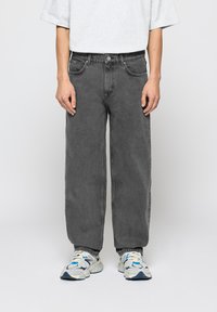 Osoba w luźnych, ciemnoszarych dżinsach oraz białych sneakersach z niebieskimi i czarnymi detalami, stojąca na jasnym, jednolitym tle.