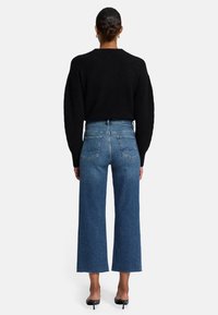 Maglione nero oversize, jeans blu a vita alta e gamba larga con orlo sfrangiato e dettagli di cucitura sulle tasche posteriori, indossati con sandali neri con tacco.