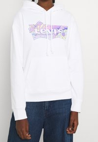 Persona vistiendo una sudadera blanca con un colorido logo de Levi's en tonos pastel, combinada con jeans de mezclilla azules, de pie contra un fondo liso.