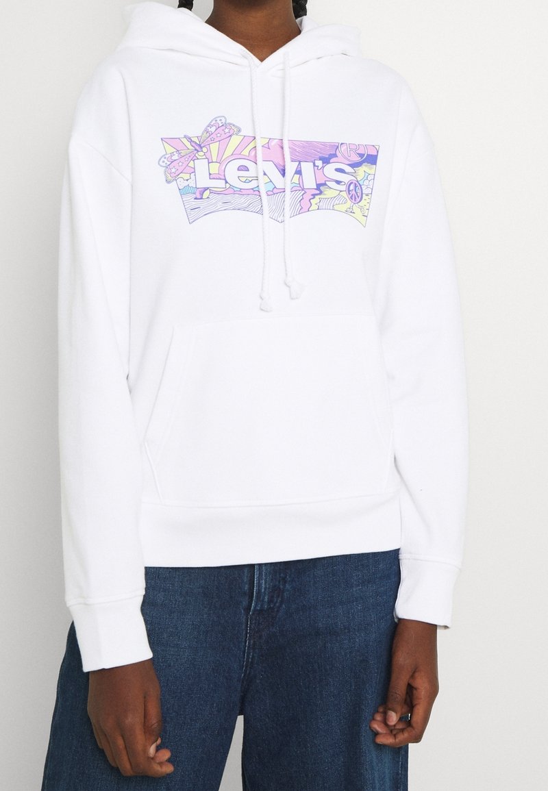 Persona vistiendo una sudadera blanca con un colorido logo de Levi's en tonos pastel, combinada con jeans de mezclilla azules, de pie contra un fondo liso.