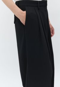 Črne nabirane hlače z gladko teksturo, z bočnimi žepi in prilagojenim pasom, prikazane s strani z roko v žepu.