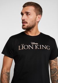 Crni pamučni T-shirt s grafikom "Kralj lavova" u zlatnom pismu. Kratke rukave i okrugli izrez. Glatka tekstura.