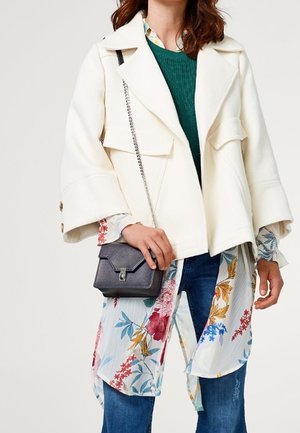 White, textured jacket with wide sleeves, layered over a floral blouse and green sweater. A small grey crossbody bag is held in one hand.